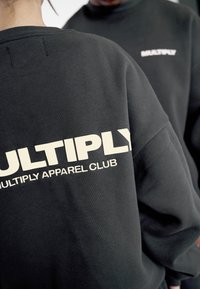 Felpa nera con un grande logo beige "MULTIPLY" e "MULTIPLY APPAREL CLUB" sul retro. Tessuto morbido con vestibilità ampia.