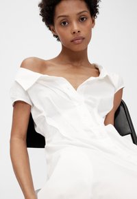 Witte overhemdjurk met korte mouwen, voorzien van een off-shoulder ontwerp, lichtgewicht stof en een losse pasvorm, zittend op een zwarte stoel.