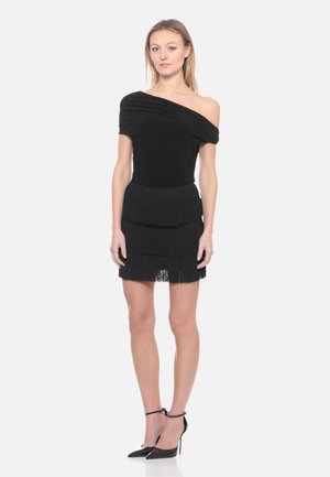 Donna che indossa un vestito nero con scollo a spalle scoperte e gonna a balze con frange, scarpe nere con tacco alto e cinturino alla caviglia, in piedi davanti a uno sfondo bianco.