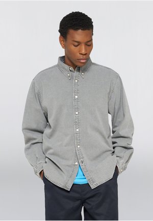 Jeune homme aux cheveux courts et bouclés, portant une chemise grise boutonnée par-dessus un t-shirt bleu, mains dans les poches, regardant vers le bas.