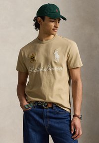 Νέος άνδρας με μπεζ μπλουζάκι Ralph Lauren με λογότυπα, μπλε τζιν, ριγέ ζώνη, πράσινο καπέλο και ρολόι χειρός, στέκεται σε απλό φόντο.