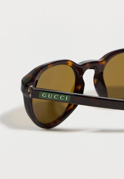 Lunettes de soleil écaille de tortue avec des verres marron, présentant une plaque logo Gucci vert foncé sur la branche.