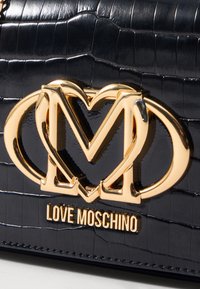 Borsa nera in eco-pelle effetto coccodrillo con logo in metallo dorato che presenta forme intrecciate di M e cuore. Texture liscia, bordi cuciti.