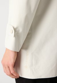 Chaqueta blanca impermeable con una textura suave, que cuenta con un puño de botón a presión y un discreto logo "GORE-TEX" cerca del dobladillo.