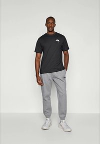 Homme debout en t-shirt noir, pantalon de jogging gris et baskets blanches sur un fond clair uni, avec une main dans la poche.