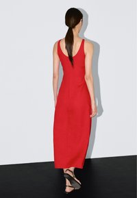 Femme aux longs cheveux bruns attachés en queue de cheval basse, portant une robe midi rouge sans manches et des sandales plates noires, debout contre un mur blanc uni.