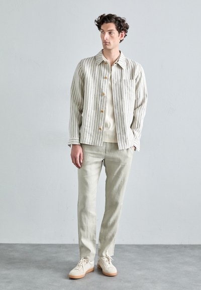 Veste légère rayée en crème et noir, associée à un pantalon beige clair et des baskets beiges. Design simple avec une coupe décontractée et une fermeture à boutons.
