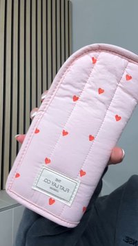 Rosa quiltad påse med små röda hjärtmönster. Har en rektangulär form och en vit etikett med varumärkesnamn längst ner.