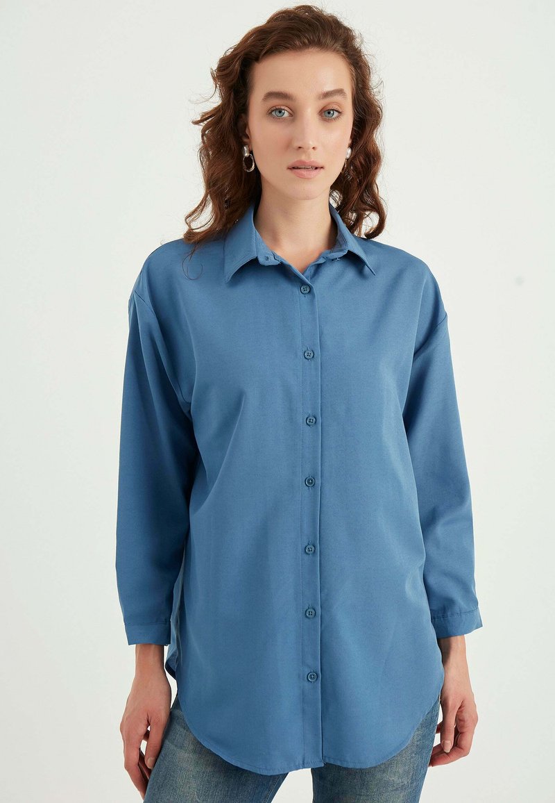 Camicia azzurra abbottonata realizzata in tessuto liscio, con colletto classico, maniche lunghe e orlo arrotondato. Sono visibili due bottoni.