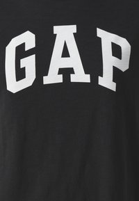 Černé bavlněné tričko s výrazným bílým nápisem "GAP". Materiál má hladkou strukturu, bez dalších vzorů nebo akcentů.