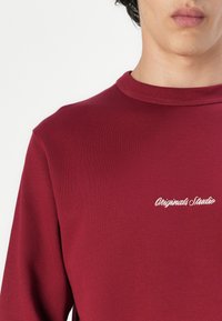 Burgundy crew neck sweatshirt made of smooth fabric, featuring white embroidered text "Originals Studio" on the front.