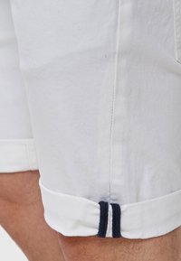 Białe, zwinięte szorty z granatowym podwójnym paskiem w okolicy mankietu na nodze osoby z widoczną skórą i włosami.