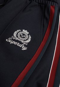 Granatowe dresy z logo. Posiadają haftowany biały napis "Superdry" oraz czerwone i zielone paski po bokach. Materiał jest gładki z lekkim połyskiem.