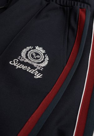 Granatowe dresy z logo. Posiadają haftowany biały napis "Superdry" oraz czerwone i zielone paski po bokach. Materiał jest gładki z lekkim połyskiem.