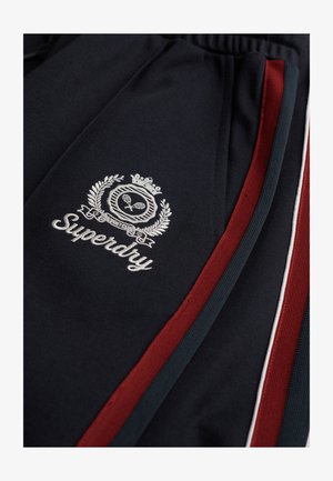 Granatowe dresy z logo. Posiadają haftowany biały napis "Superdry" oraz czerwone i zielone paski po bokach. Materiał jest gładki z lekkim połyskiem.