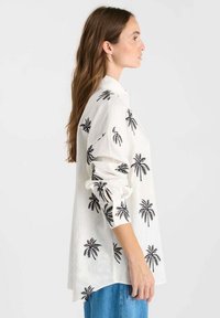 White linen shirt featuring black palm tree prints, long sleeves, a button-down collar, and a relaxed fit with a longer back hem.