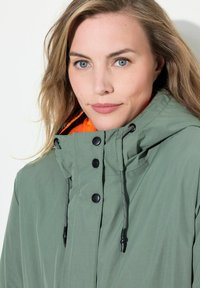 Grön huva jacka med en texturerad yta, prydd med svarta tryckknappar, justerbara snören och en orange innerfoder.