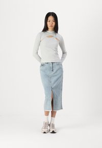Svetlo siva majica s dolgimi rokavi in izrezom, v kombinaciji z-denim midi krilom z razporkom spredaj. Manekenka nosi svetle superge in nogavice.
