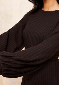 Robe noire côtelée avec manches longues et volumineuses. Le tissu texturé met en valeur les lignes verticales. Encolure arrondie et corsage ajusté.