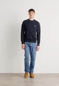 Sudadera navy con puños y dobladillo acanalados, combinada con jeans azules claros y zapatos marrones. El modelo está de pie frente a un fondo blanco liso.