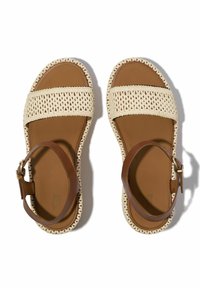 Sandalias marrones con una correa trenzada en color crema, cierre en el tobillo ajustable y una suela texturizada. Diseño de punta abierta con tacón plano.
