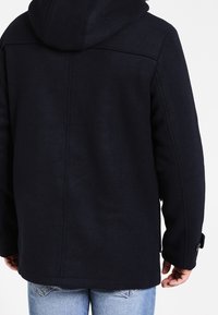 Persona che indossa un cappotto con cappuccio nero e jeans azzurri chiari visto da dietro su uno sfondo bianco semplice.