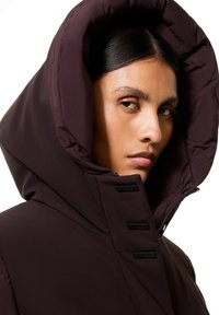 Giacca puffer bordeaux con un grande cappuccio, dotata di una chiusura frontale con zip e dettagli testurizzati al colletto. Tessuto morbido e liscio con un design elegante.