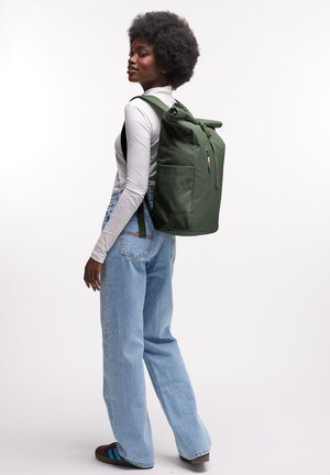 Donna con camicia bianca a maniche lunghe e jeans blu che indossa un grande zaino verde con chiusura a rotolo, in piedi contro uno sfondo chiaro e uniforme.