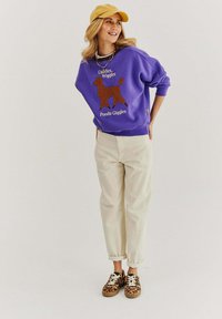 Fialová oversized mikina s potiskem "Poodle Giggles", béžové kalhoty s manžetami a tenisky s leopardím potiskem. Doplněno žlutou kšiltovkou.