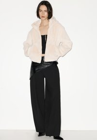 Kremasta jakna od umjetnog krzna s kapuljačom, kratkog kroja, s patentnim zatvaračem sprijeda i bočnim džepovima, u kombinaciji s crnim hlačama širokih nogavica i crnim topom.