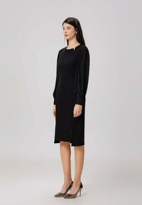 Robe noire midi avec de longues manches bouffantes, un décolleté carré et une fente sur le côté. Fabriquée en tissu texturé, associée à des escarpins à bout pointu.