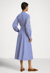 Mulher usando um vestido midi de manga comprida com riscas verticais azuis e brancas, com detalhes em renda na manga, combinado com sapatos planos castanhos.