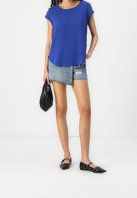 Haut bleu à manches courtes, associé à une mini-jupe en denim avec ourlet effiloché, et des ballerines noires à œillets. Tenant un sac à main noir.