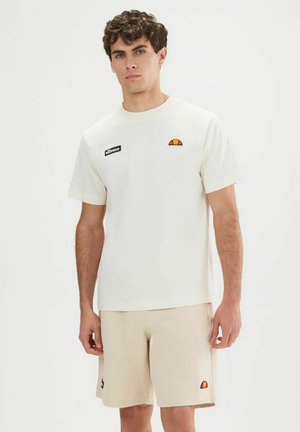 Jeune homme portant un t-shirt ellesse crème et un short assorti, debout devant un fond blanc uni.