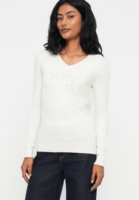 Maglione bianco a maniche lunghe a V realizzato in morbido materiale, con la scritta "Guess est 1981" in un design leggermente in rilievo sul fronte.