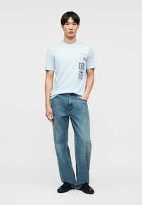 Άνδρας που στέκεται με το ένα χέρι στην τσέπη, φοράει ανοιχτό μπλε μπλουζάκι με γραφικό σχέδιο, πλατύπεδη μπλε τζιν και μαύρα παπούτσια σε μονόχρωμο φόντο.