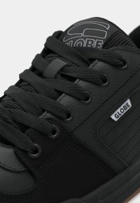 Fekete sneaker texturált bőr és velúr felsőrésszel, lapos fekete fűzővel, párnázott nyelvvel, rajta logóval, és gumitalppal.