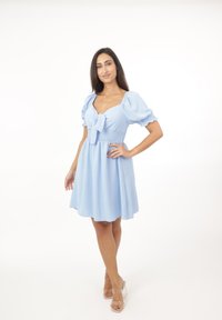 Hellblaues Kleid mit Puffärmeln, gerafftem Bund und einer Frontbindeschleife; knielang mit strukturiertem Stoff. Getragen mit transparenten Blockabsatzsandalen.