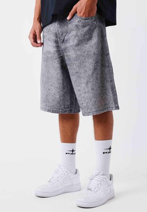 Man draagt grijze, gespikkelde knielange shorts, witte sokken met zwart logo en witte sneakers tegen een effen achtergrond.