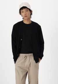 Svart cardigan med en avslappet passform, med fem knapper og ribbestrikkede mansjetter. Kombinert med en svart skjorte og beige bukser med snøring.