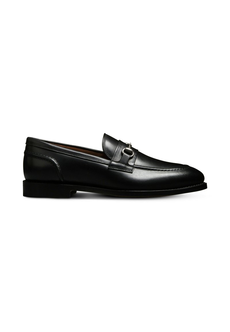 Mocassin noir en cuir à enfiler avec semelle plate, arborant une boucle en métal argenté décorative sur le dessus et des détails de couture visibles.