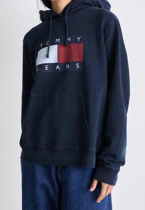 Person trägt einen marineblauen Tommy Jeans Hoodie mit einer Fronttasche und blaue Jeans, dargestellt vom Hals bis zur Hüfte vor einem einfarbigen Hintergrund.