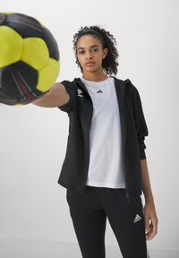 Mujer joven con chándal negro de Adidas y camiseta blanca extiende la mano sosteniendo una pelota de balonmano amarilla y negra hacia la cámara.