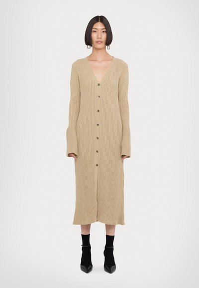 Vestido de punto acanalado beige con mangas largas acampanadas, escote en V profundo y botones en la parte delantera. Silueta hasta la rodilla, combinado con botas negras de tobillo.