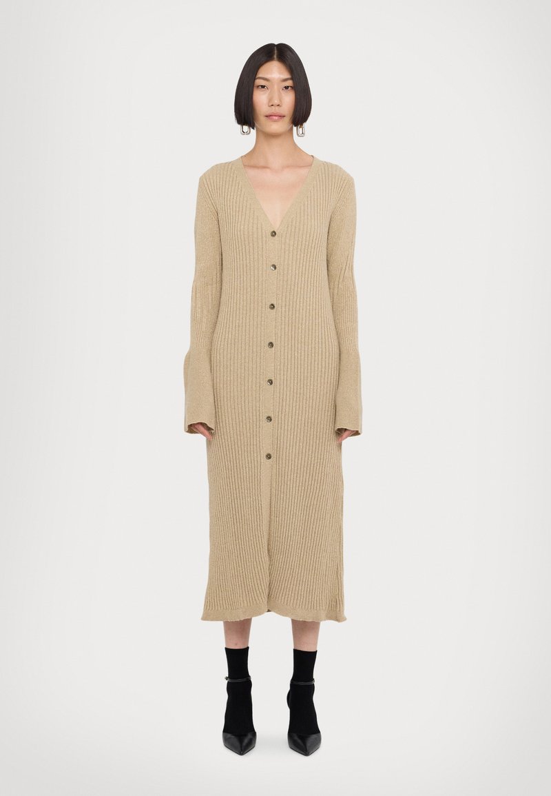 Beige geribbelde gebreide jurk met lange klokmouwen, diepe V-hals en knopen aan de voorkant. Knelengte silhouet, gecombineerd met zwarte enkellaarsjes.