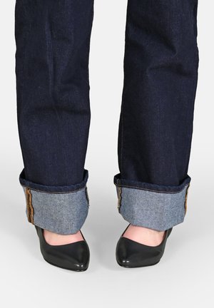Jean en denim bleu marine avec des poignets retroussés révélant un tissu en denim plus clair en dessous, associés à des talons noirs pointus.