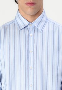 Jasnoniebieska koszula z guzikami, w pionowe białe i granatowe paski, z klasycznym kołnierzykiem i pięcioma białymi guzikami z przodu.