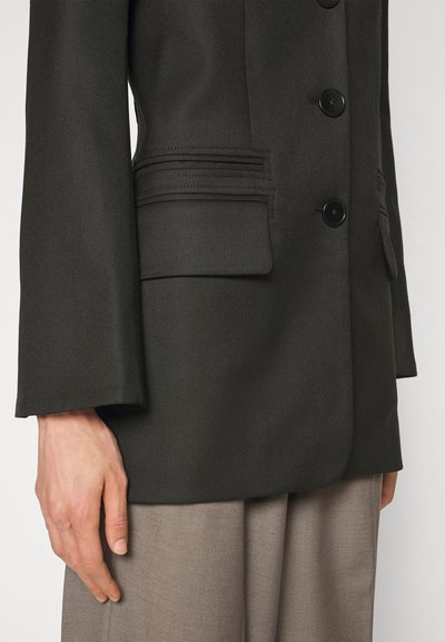 Zwarte op maat gemaakte blazer met een gestructureerd ontwerp, knoopsluiting en dubbele zakdetails, gecombineerd met lichtgekleurde geplooide broeken.