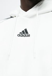 Biela mikina s kapucňou s čiernym vyšívaným logom Adidas na hrudi, so sťahovacími šnúrkami a jemnou textúrou.