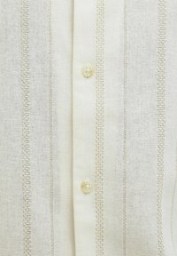 Chemise de couleur crème avec des rayures verticales et des coutures décoratives. Elle a une fermeture à boutons avec une finition lisse et brillante.